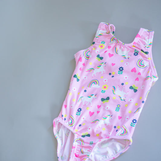 Pink unicorn and rainbow print gymnastics leotard for girls by cbcsportswear. Features colorful hearts, flowers, and bows. Made from soft, comfortable fabric, perfect for young gymnasts.